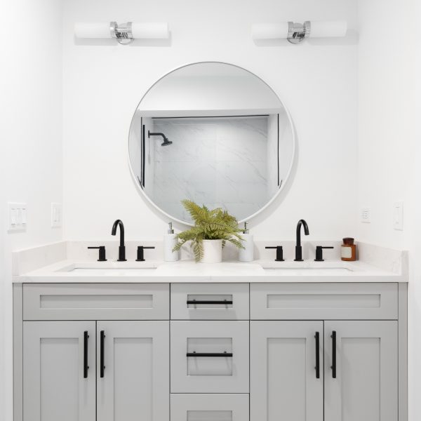 A renovated bathroom with a grey vanity cabinet, circular mirror with a view to a shower, and back faucets.
